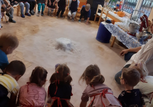 Dzieci siedzą na krzesłach ustawionych w półkole. Na środku na brązowej podłodze widać dym wytwarzany za pomocą suchego lodu.