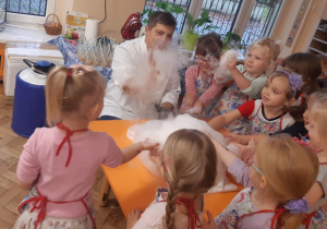 Dziewczynki ustawione wokół pomarańczowego stolika dotykają piany utworzonej z wody, suchego lodu i płynu do naczyń.