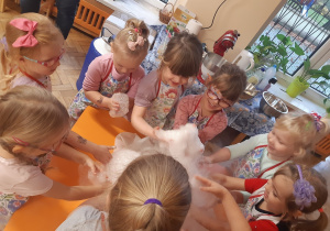 Dziewczynki ustawione wokół pomarańczowego stolika dotykają piany utworzonej z wody, suchego lodu i płynu do naczyń.