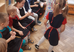 Dwie dziewczynki ubrane w kolorowe fartuszki rozdają dzieciom siedzącym na krzesełkach soczki nalane w przezroczyste kubeczki ze słomką.