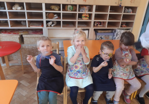 Dwie dziewczynki w kolorowych fartuszkach i dwóch chłopców w czarnych fartuszkach siedzą na krzesełkach. Z ust dzieci wydobywa się dym.