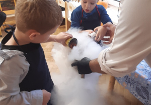 Dwóch chłopców wlewa zawartość metalowych puszek do metalowej miski. Z miski wydobywa się biały dym.