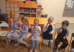 Trzy dziewczynki i dwóch chłopców siedzi na krzesełkach i je lody. W tle widać pomarańczowe stoliki i szafkę.