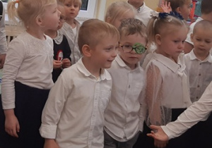 Dzieci ubrane są na galowo. Stoją na kolorowym dywanie. Nad dziećmi znajduje się patriotyczna girlanda.