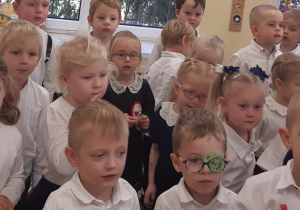 Dzieci ubrane są na galowo. Stoją na kolorowym dywanie. Nad dziećmi znajduje się patriotyczna girlanda.