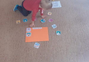 Chłopiec klęczy na beżowym dywanie, na którym leży pomarańczowy karton oraz kolorowe obrazki przedstawiające prawa dziecka.