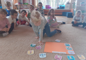 Dziewczynka klęczy na beżowym dywanie, na którym leży pomarańczowy karton oraz kolorowe obrazki przedstawiające prawa dziecka. W tle widać inne dzieci siedzące na dywanie.