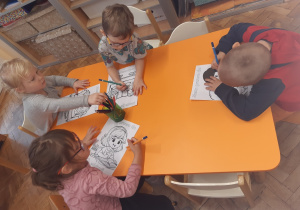 Dwie dziewczynki i dwóch chłopców siedzi przy pomarańczowym stole i kolorują kredkami obrazki.