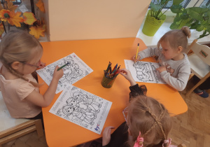 Trzy dziewczynki siedzą przy pomarańczowym stole i kolorują kredkami obrazki.