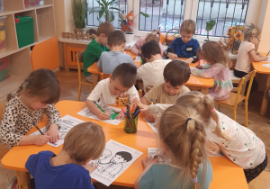 Dzieci siedzą przy pomarańczowych stołach i kolorują kredkami obrazki przedstawiające prawa dziecka.