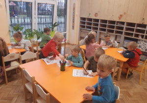 Dzieci siedzą przy pomarańczowych stołach i kolorują kredkami obrazki przedstawiające prawa dziecka.