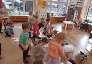 Dzieci chodzą po beżowym dywanie, na którym rozłożone są białe kartki papieru. Dzieci rozdają czerwone papierowe serduszka swoim rówieśnikom.