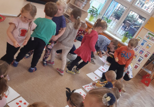 Dzieci chodzą po beżowym dywanie, na którym rozłożone są białe kartki papieru. Dzieci rozdają czerwone papierowe serduszka swoim rówieśnikom.