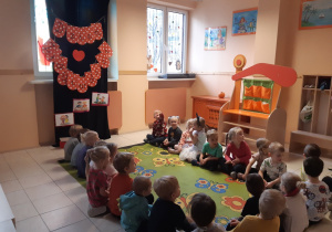 Dzieci siedzą na zielonym dywanie w kolorowe kwiaty i motyle. Na oknie powieszony jest czarny materiał, na którym umieszczony jest napis na czerwonych kartkach z białymi kropkami oraz trzy ilustracje.