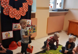 Chłopiec stoi na beżowych kafelkach, w rękach trzyma kolorową ilustrację. Przed chłopcem na zielonym dywanie w kolorowe kwiaty i motyle siedzą dzieci.