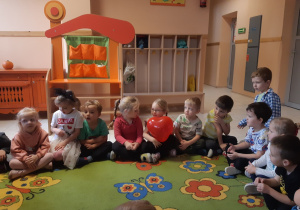 Dzieci siedzą w kole na zielonym dywanie w kolorowe kwiaty i motyle. Dziewczynka w okularach trzyma w rękach czerwony balon w kształcie serca.