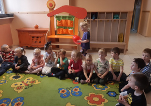 Dzieci siedzą w kole na zielonym dywanie w kolorowe kwiaty i motyle. Jedna dziewczynka idzie z czerwonym balonem w kształcie serca za plecami dzieci.