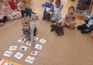 Dzieci siedzą na brzegu beżowego dywanu. W rękach trzymają swoje pluszowe misie. Jeden chłopiec klęczy na środku dywanu koło ilustracji przedstawiających bohaterów bajek o misiach.