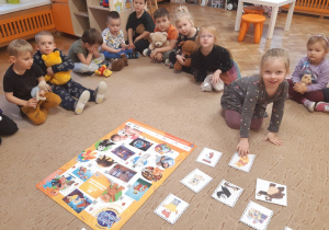 Dzieci siedzą na brzegu beżowego dywanu. W rękach trzymają swoje pluszowe misie. Jedna dziewczynka klęczy na środku dywanu obok ilustracji przedstawiających bohaterów bajek o misiach.