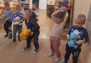 Dzieci stoją na beżowym dywanie, w rękach trzymają pluszowe misie.