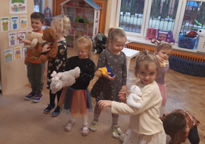 Dzieci stoją na beżowym dywanie. Dzieci mają położone na rękach pluszowe misie.