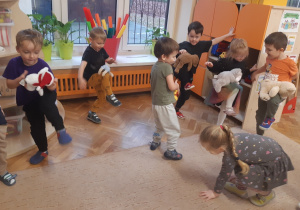 Dzieci stoją na jednej nodze. Na nodze uniesionej w górze mają położonego pluszowego misia.