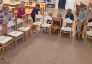 Dzieci znajdują się za krzesełkami ustawionymi na brzegu beżowego dywanu. Dzieci chowają pluszowe misie pod krzesłami.