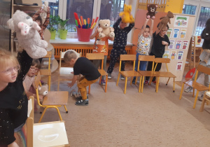 Dzieci znajdują się za krzesełkami ustawionymi na brzegu beżowego dywanu. Dzieci trzymają pluszowe misie nad krzesłami.