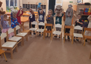 Dzieci znajdują się za krzesełkami ustawionymi na brzegu beżowego dywanu. Dzieci trzymają pluszowe misie nad krzesłami.