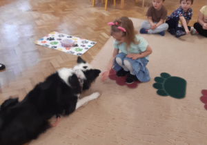 Dziewczynka kuca przed biało-czarnym psem i podaje mu na ręku psi przysmak. W tle widać dzieci siedzące na beżowym dywanie.