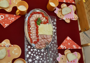 Na zdjęciu widoczny jest świątecznie nakryty stół. Na półmisku ułożone są pomidory koktajlowe, wędlina i ser żółty tworzące kształt cukrowej laski.