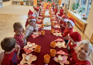 Dzieci siedzą przy długim stole przykrytym czerwonym obrusem, na którym położony jest biały bieżnik w srebrne gwiazdki oraz papierowe talerzyki w kształcie ludzików w piernika, biało-czerwone słomki i pomarańczowe plastikowe kubki. Dzieci ubrane są na czerwono i mają na głowach czerwone mikołajkowe czapki.