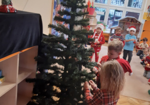 Na zdjęciu widoczna zielona choinka, przy której stoi troje dzieci.