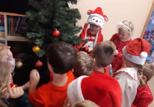 Dzieci w czerwonych strojach i czerwonych czapkach na głowie stoją wokół zielonej choinki.