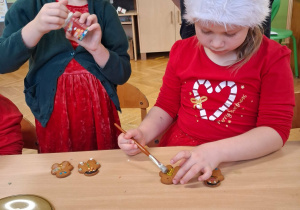 Na zdjęciu dzieci ubrane na czerwono jedno z dzieci w mikołajowej czapce, dzieci ozdabiają pierniki