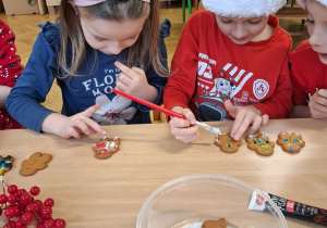 Na zdjęciu dzieci ubrane na czerwono jedno z dzieci w mikołajowej czapce, dzieci ozdabiają pierniki