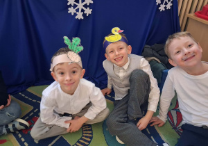 Na zdjęciu troje dzieci z grupy zielonej podczas przygotowania do przedstaiwnie z okazji "Dnia Babci i Dziadka", dzieci mają na sobie opaski z postaciami z wiersza Jana Brzechwy "Rzepka"