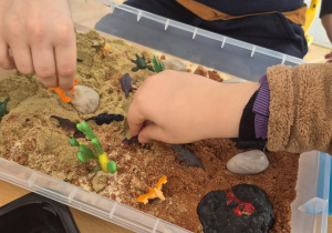 Na zdjeciu pojemnik z ziemią w któej ukrytę są dinzoaury, dzieci poszukują dinozaurów