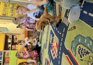 Na zdjęciu dzieci siedzą na dywanie wraz ze swoimi dinozaurami, na dywanie jest brązowy duży dinozaur