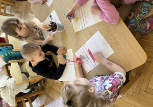 Na zdjeciu dzieci siedzą przy stole i kolorują opaskę na głowę z motywem dinozaurów