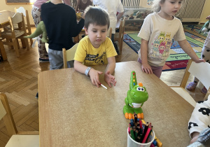 Na zdjeciu dzieci siedzą przy stole na stole jest zielony dinozaur