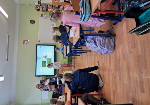 Na zdjęciu dzieci siedzące w ławkach w szkole na wprost jest tablica multimedialna