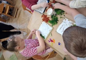 Dzieci pracują przy stołach na stole leżą zielone materiały dzieci przyjklejają je do tacek