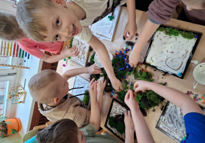 Dzieci pracują przy stołach na stole leżą zielone materiały dzieci przyjklejają je do tacek