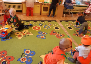 Dzieci z grupy żółtej w strojach jesiennych zwierząt bawią się na dywanie autkami.