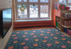 Na zdjęciu grupa pomarańczowa, na fotografii widać dywan na podłodze na ścianie obok wisi sprzęt multimedialny na wprost widać okno, z prawej półkę z zabawkami