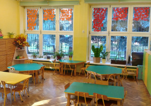Na zdjęciu sala grupy żółtej na zdjęciu widoczne 4 stoły w kolorach zielonym i żółtym oraz okna przyozdobione dekoracjami