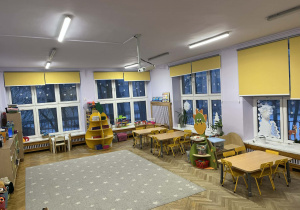 Na fotografii grupa fioletowa, na środku sali leży dywan, po lewej stronie tablica multimedialna po prawej stronie stoliki i krzesła dla dzieci, na wprost kąciki tematyczne dla dzieci