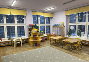 Na fotografii grupa fioletowa, na środku sali leży dywan, po lewej stronie tablica multimedialna po prawej stronie stoliki i krzesła dla dzieci, na wprost kąciki tematyczne dla dzieci