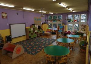 Na fotografii grupa fioletowa, na środku sali leży dywan w sowy, po lewej stronie tablica multimedialna po prawej stronie stoliki i krzesła dla dzieci, na wprost kąciki tematyczne dla dzieci